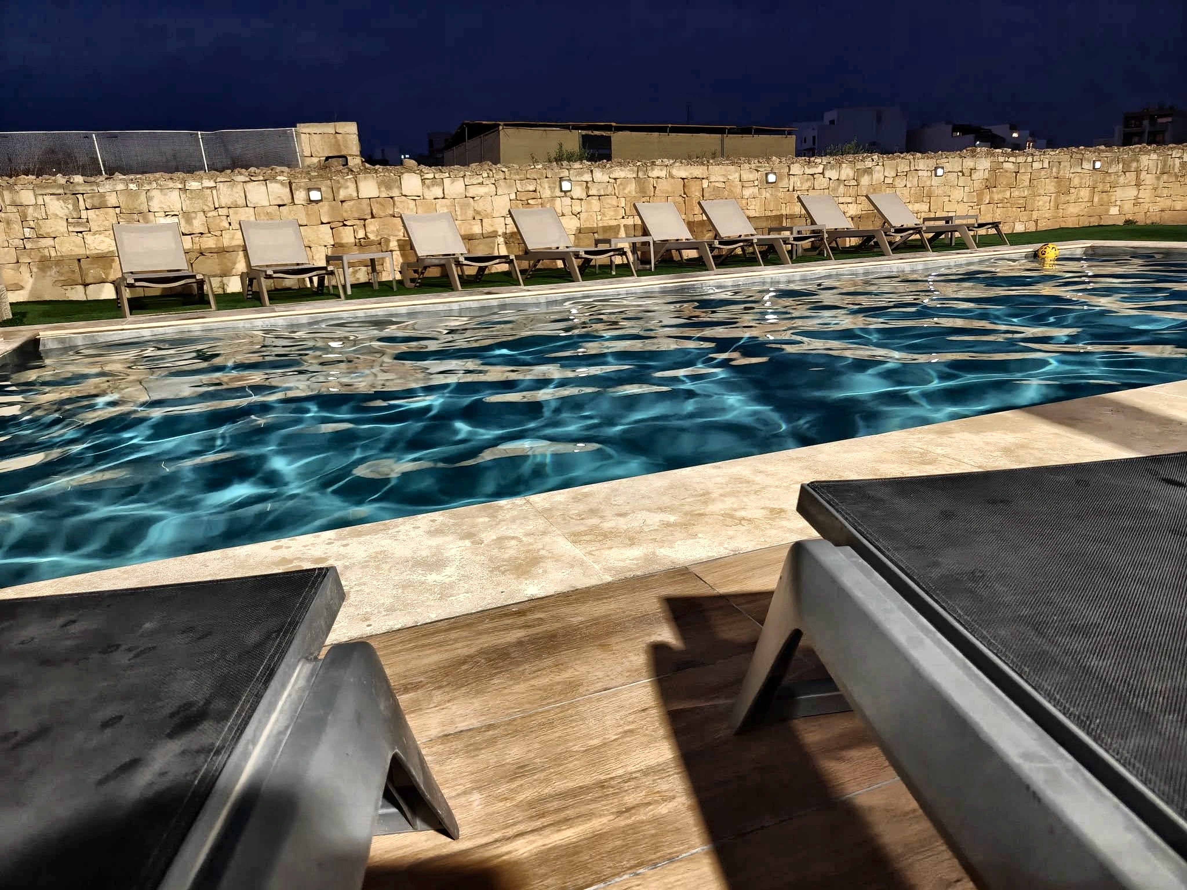 Illuminated evening private swimming pool and loungers against limestone boundary